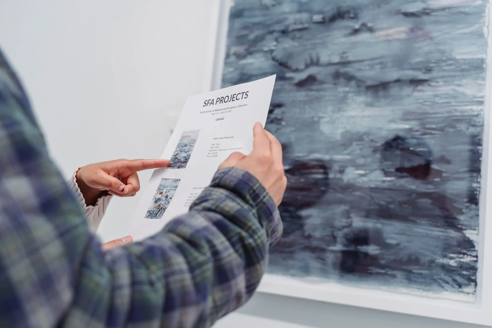 A person examining an art gallery document in front of hanging artwork