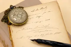 Antique pocket watch resting on a vintage journal with handwritten pages and a quill